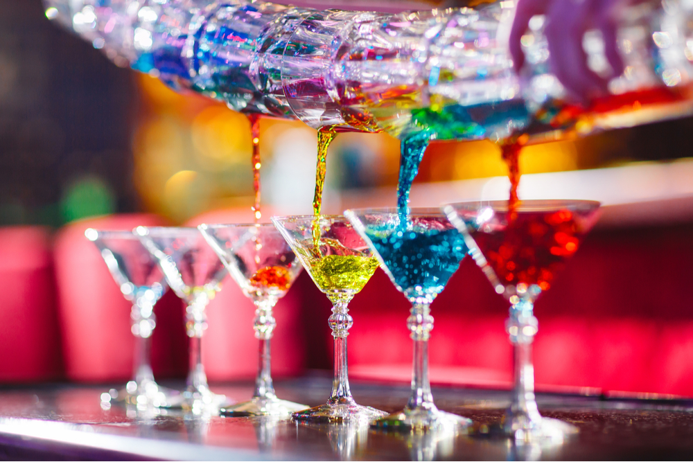 A line of martini glasses being filled with different color cocktails on a bar by a flair bartender.