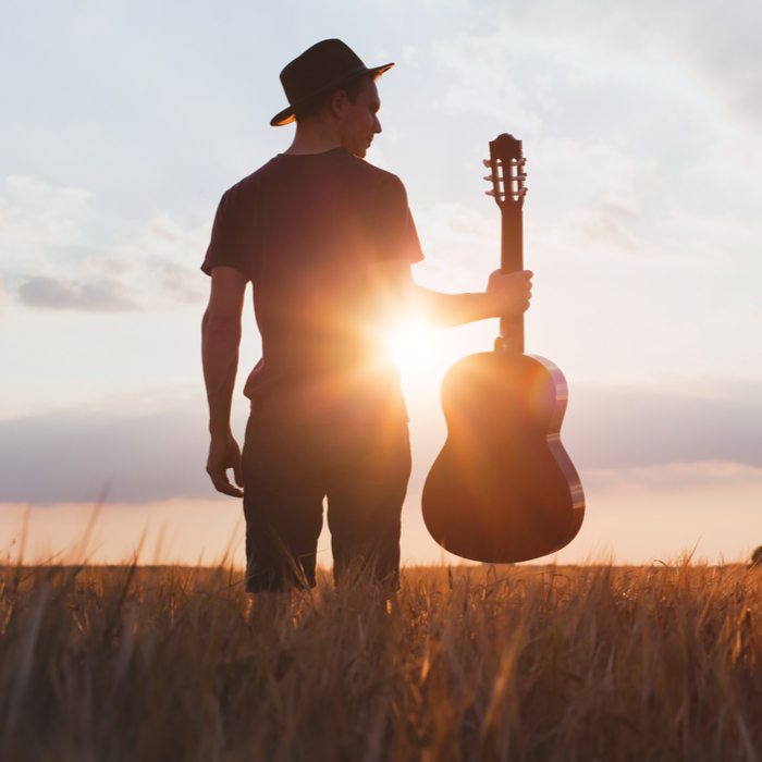 Man with a hat holding a guitar at sunset in a field on Get Out Your Guitar Day.
