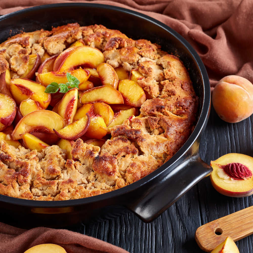 Peach cobbler in a cast iron pan. A photo of a peach cobbler recipe made in a cast iron pan, with a red cloth and peaches styled around it.