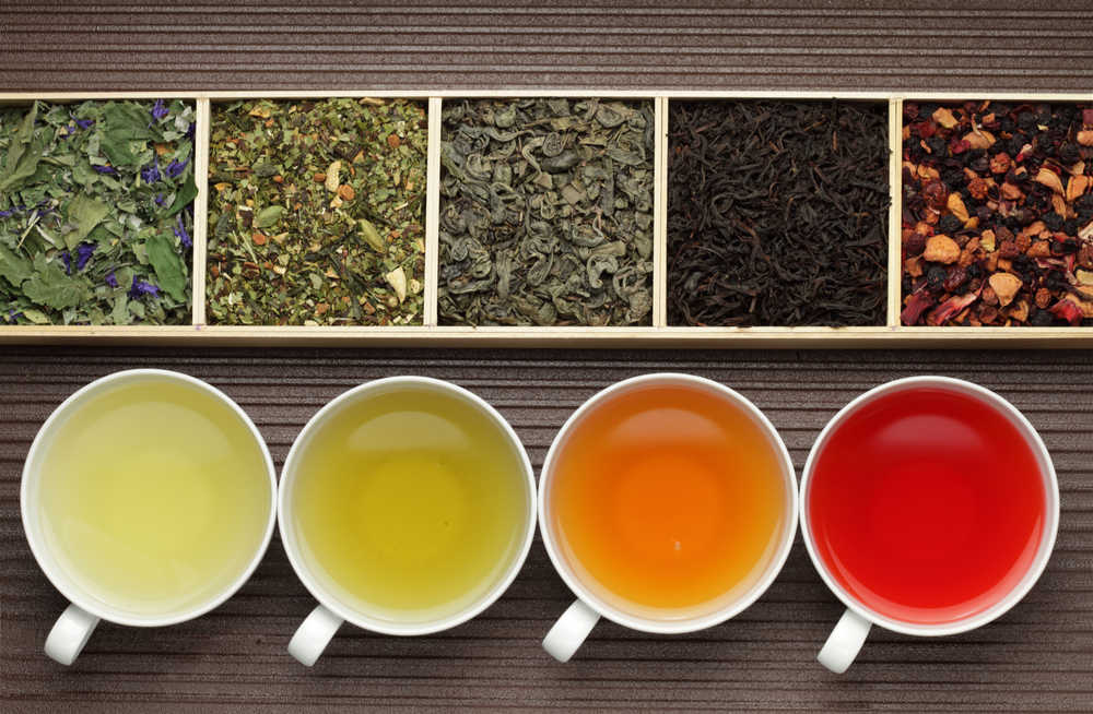Four different cups of tea from light to dark in a row below a box with five compartments each filled with a different loose leaf tea.