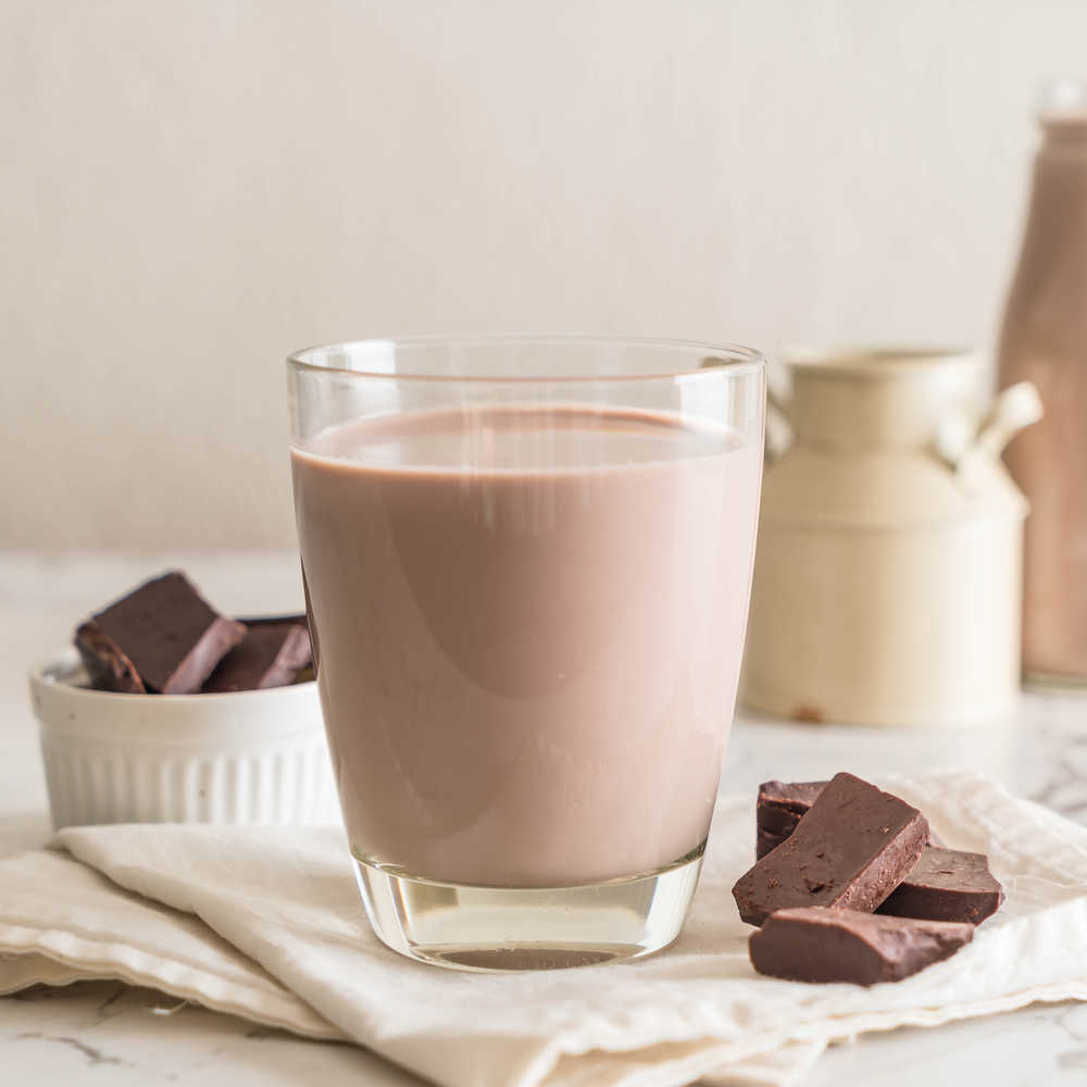 A short glass of chocolate milk on a table with linens, chocolate and kitchen props styled around it.