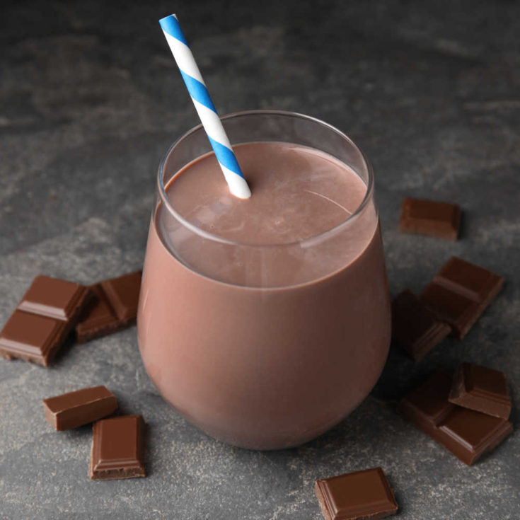 A glass of homemade chocolate milk on a slate background surrounded by chocolate squares.