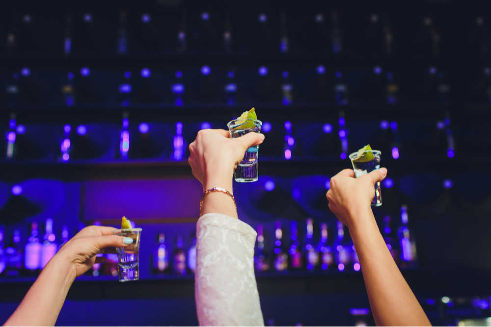 Three people cheers with shots at bar. Three hands holding shots of vodka with limes balanced on top up to the sky in a cheers motion in front of a bar with purple lighting.
