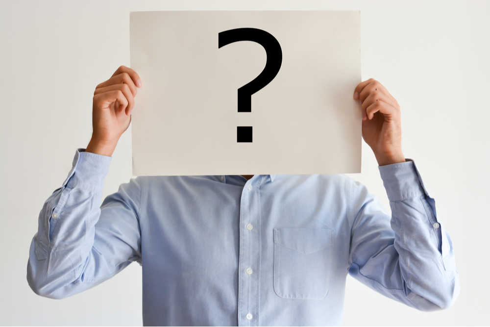 A man holding a question mark. A man in a blue button down shirt holding a white piece of paper in front of his face with a question mark on it.