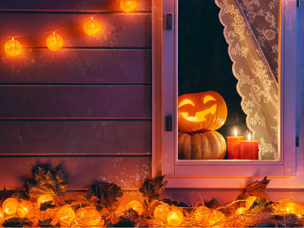 A window with a jack-o-lantern peeking out from behind a lace curtain, with leaves outside the window, and a Halloween themed set of twinkle lights.