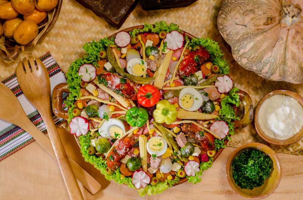 A table with wooden salad utensils, napkins, salt, spices, and potatoes next to the Guatemalan dish Fiambre, typically served on All Saints Day.