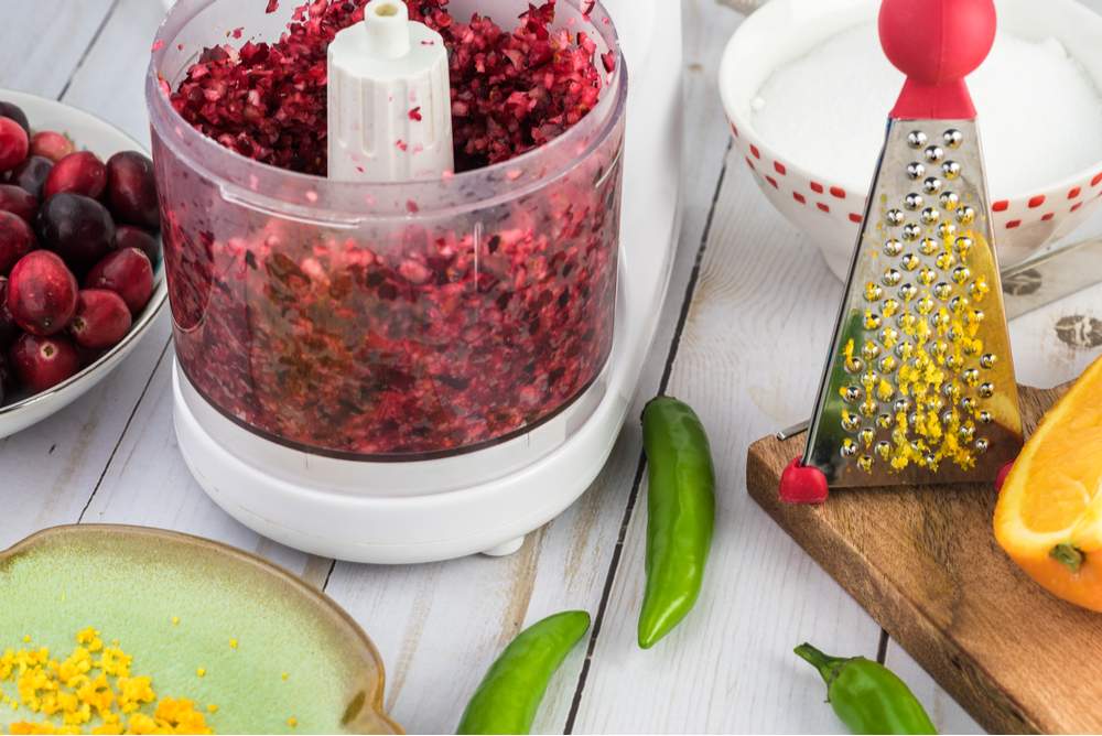 Cranberry relish in a food processor. A food processor filled with fresh cranberry relish on a white wooden table next to a grated orange, some raw cranberries and jalapenos.
