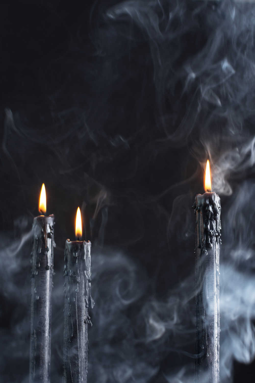 Three black candles lit against a black background with smoke and fog in the foreground.