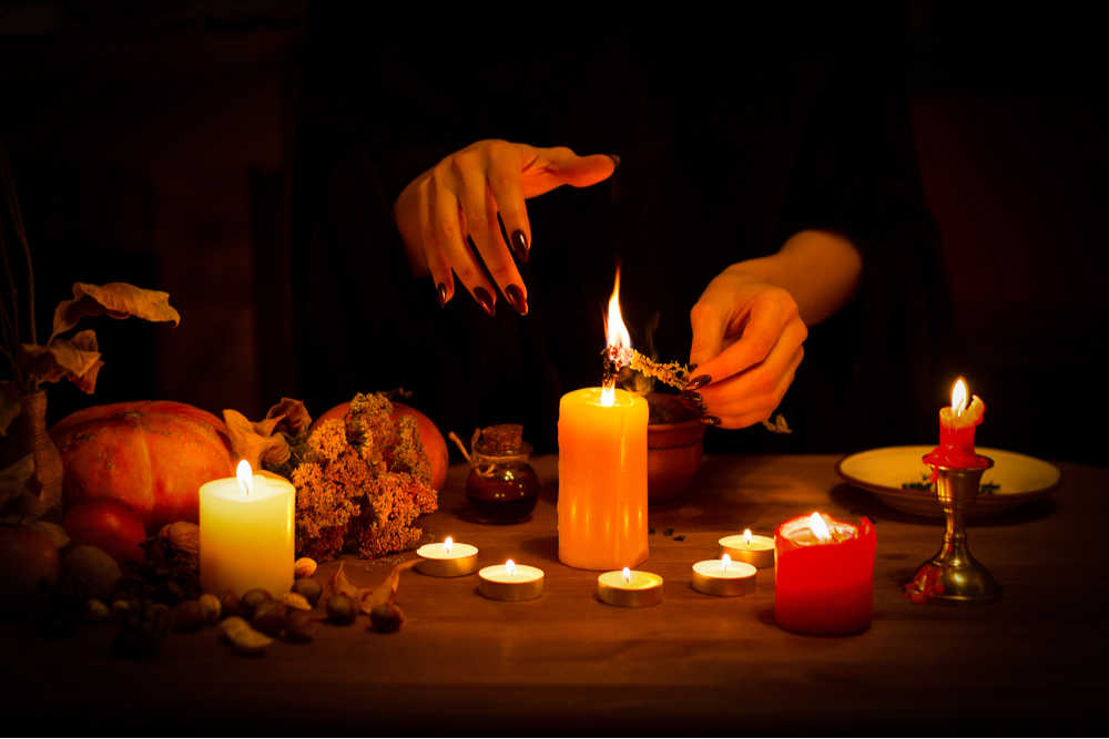 A woman with black nails lighting a large candle in the center of a semi circle of smaller lit candles as part of a Halloween tradition.