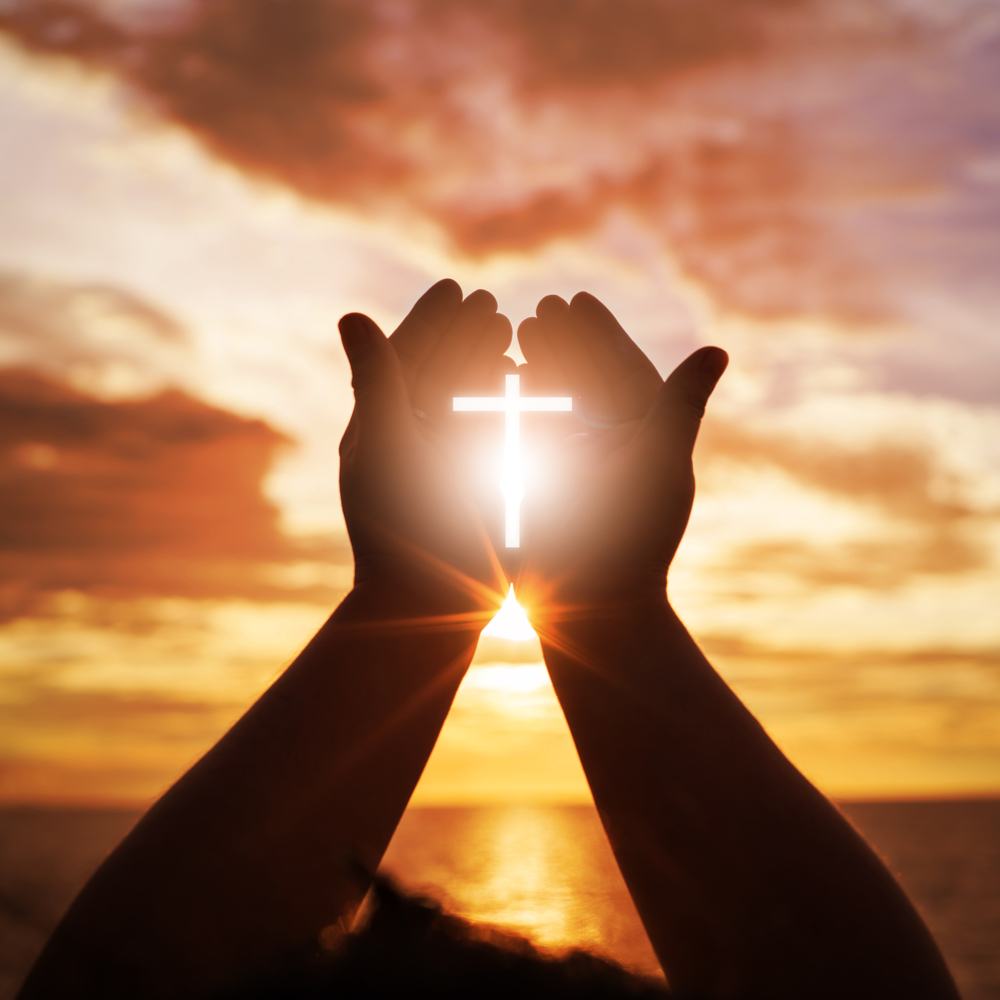 Two hands up towards an orange and yellow sky, holding a cross made out of bright white light.