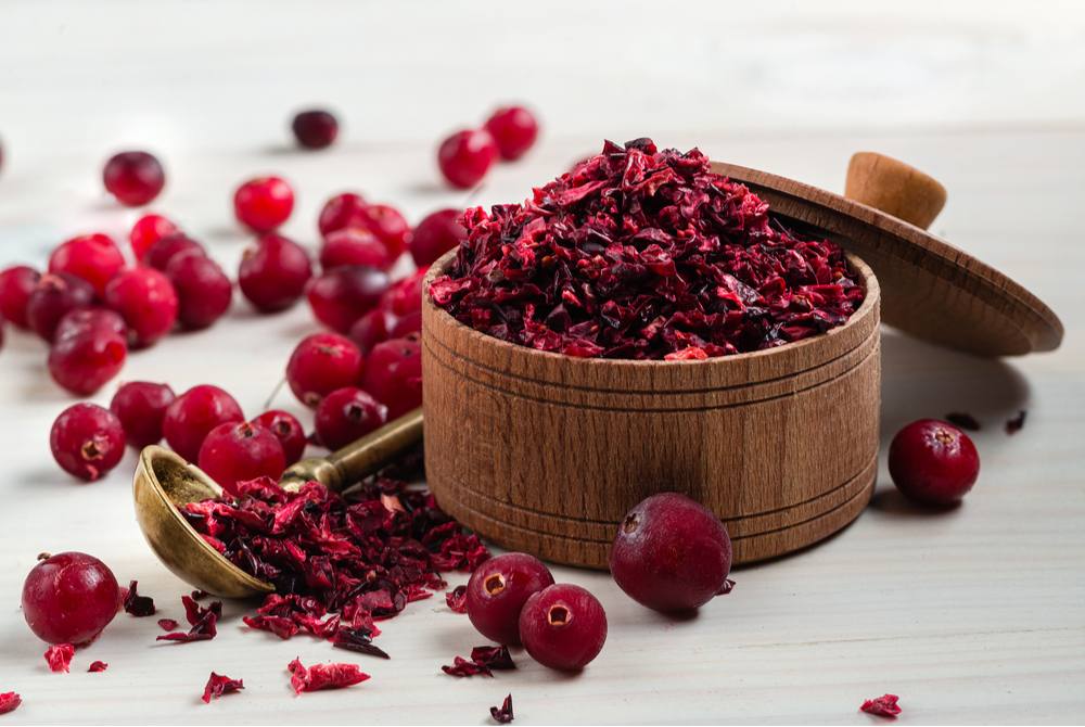 Raw cranberries and dried cranberries. A wooden container of dried cranberries next to a bunch of raw cranberries on a table, to illustrate the answer to the question "can you eat raw cranberries?".