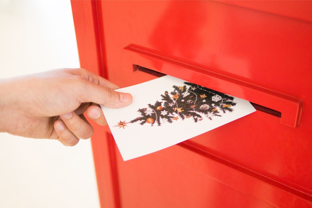 A hand putting a Christmas card with a Christmas tree in a big red mailbox to celebrate Christmas Card Day.