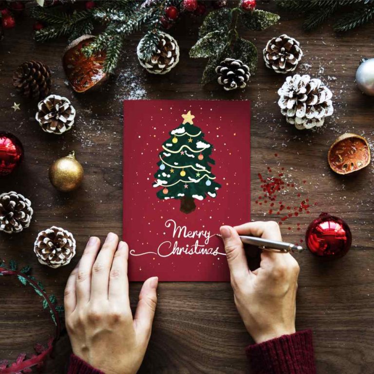 A person writing "Merry Christmas" on a red Christmas card with a tree on it, surrounded by pinecones and ornaments on Christmas Card Day.