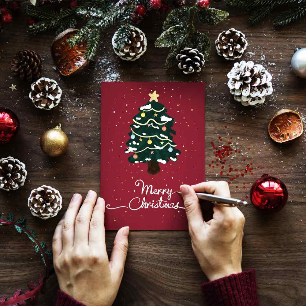 A person writing "Merry Christmas" on a red Christmas card with a tree on it, surrounded by pinecones and ornaments on Christmas Card Day.