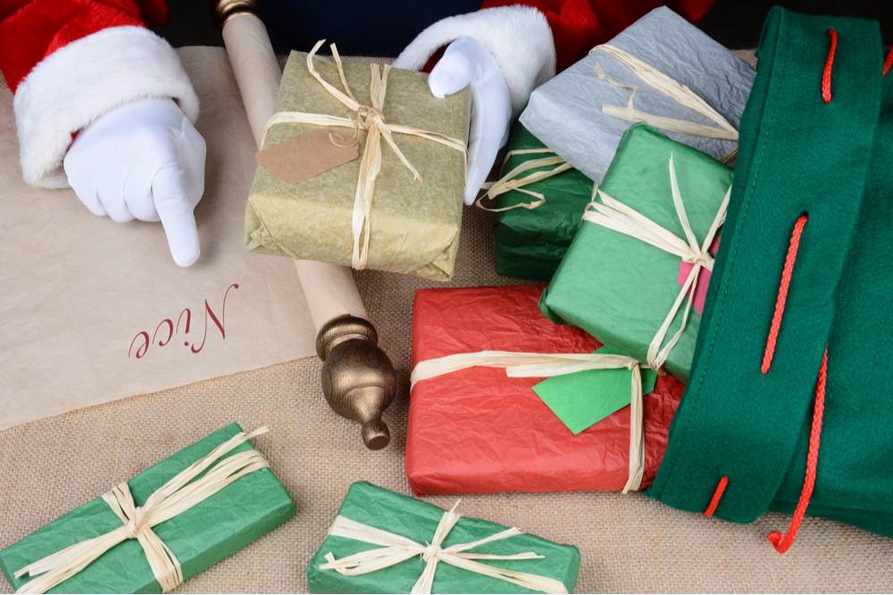 Santa with the nice list and a sack of presents. Santa pointing to the nice list holding a present that is spilling out of a sack of presents on a table.