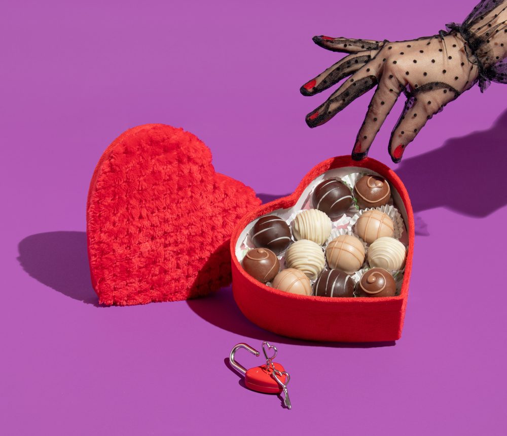 A hand in a lace glove reaching into a box of Valentine's Day chocolates and choosing a milk chocolate truffle.