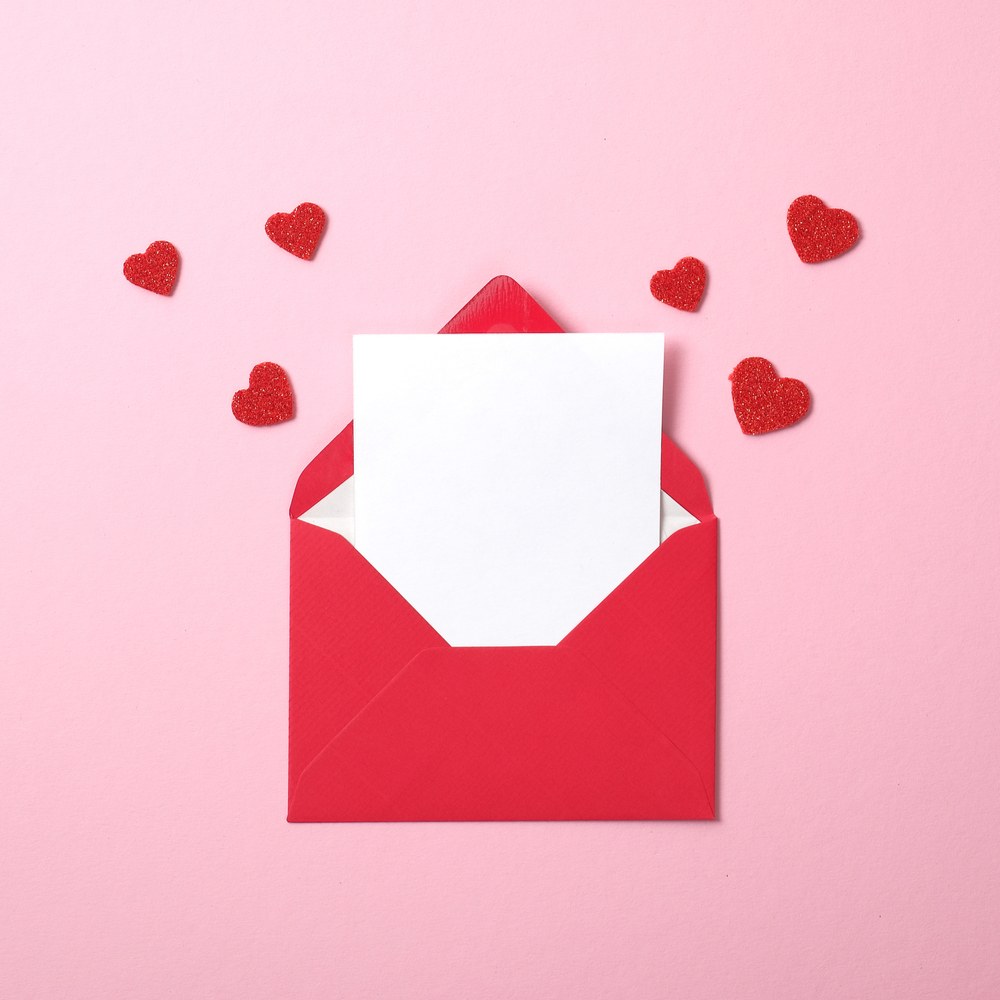 A red envelope with a love note coming out of it and little hearts around it on a pink background to symbolize early Valentine's Day cards.