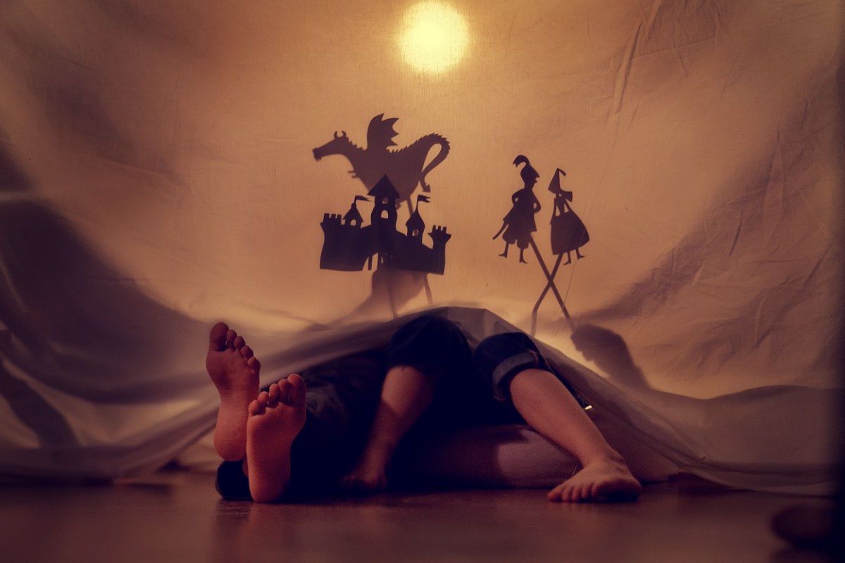 Shadow puppets A parent and child behind a sheet with their feet sticking out of it, playing with shadow puppets, including a castle, a dragon, a prince and princes to retell a Greek mythology love story and fairy tale.