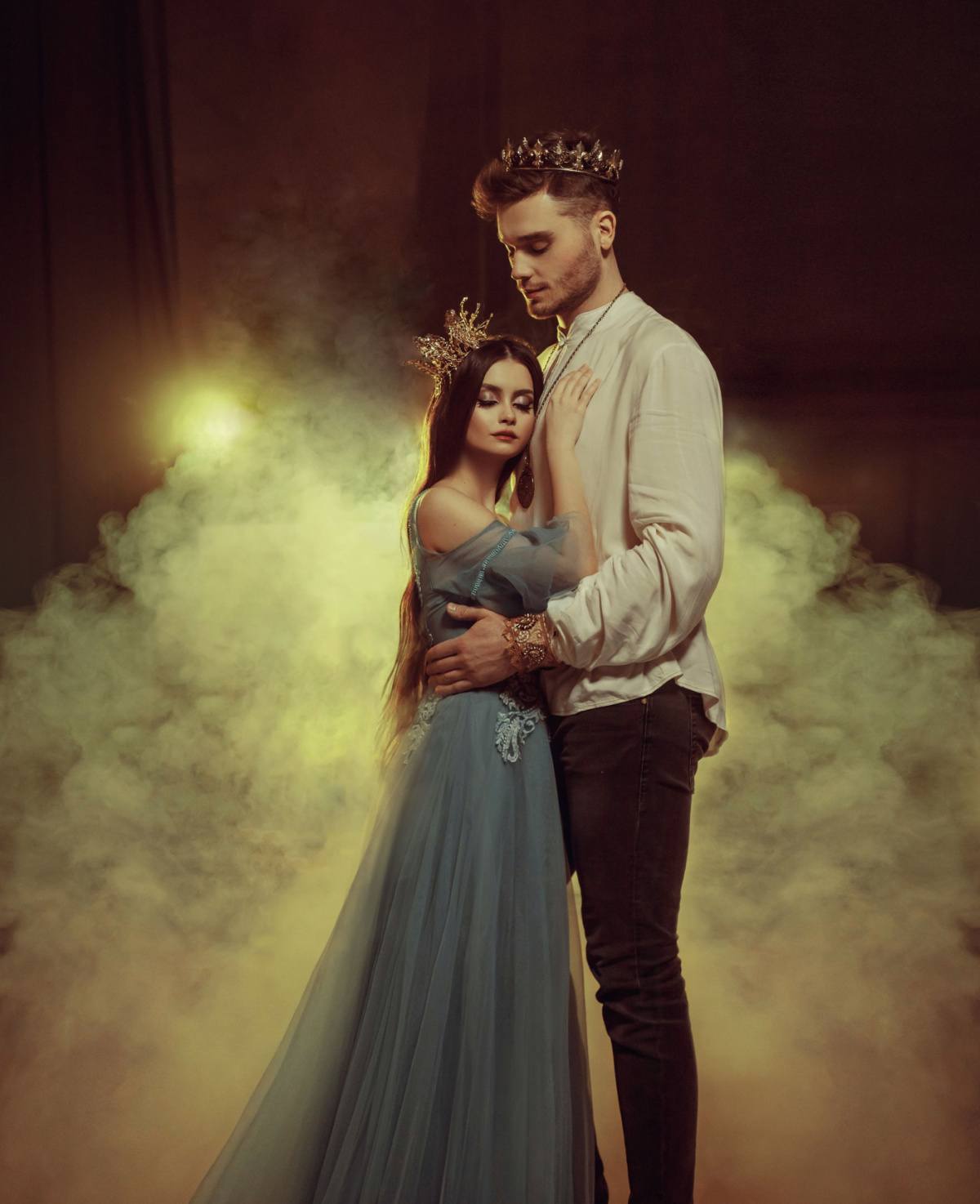 Prince and princess A moment that looks like it was taken out of a fairy tale love story, where a woman is dressed like a princess wearing a blue gown and gold crown, leaning on a man dressed like a prince wearing a gold crown, white button down shirt and black slacks.