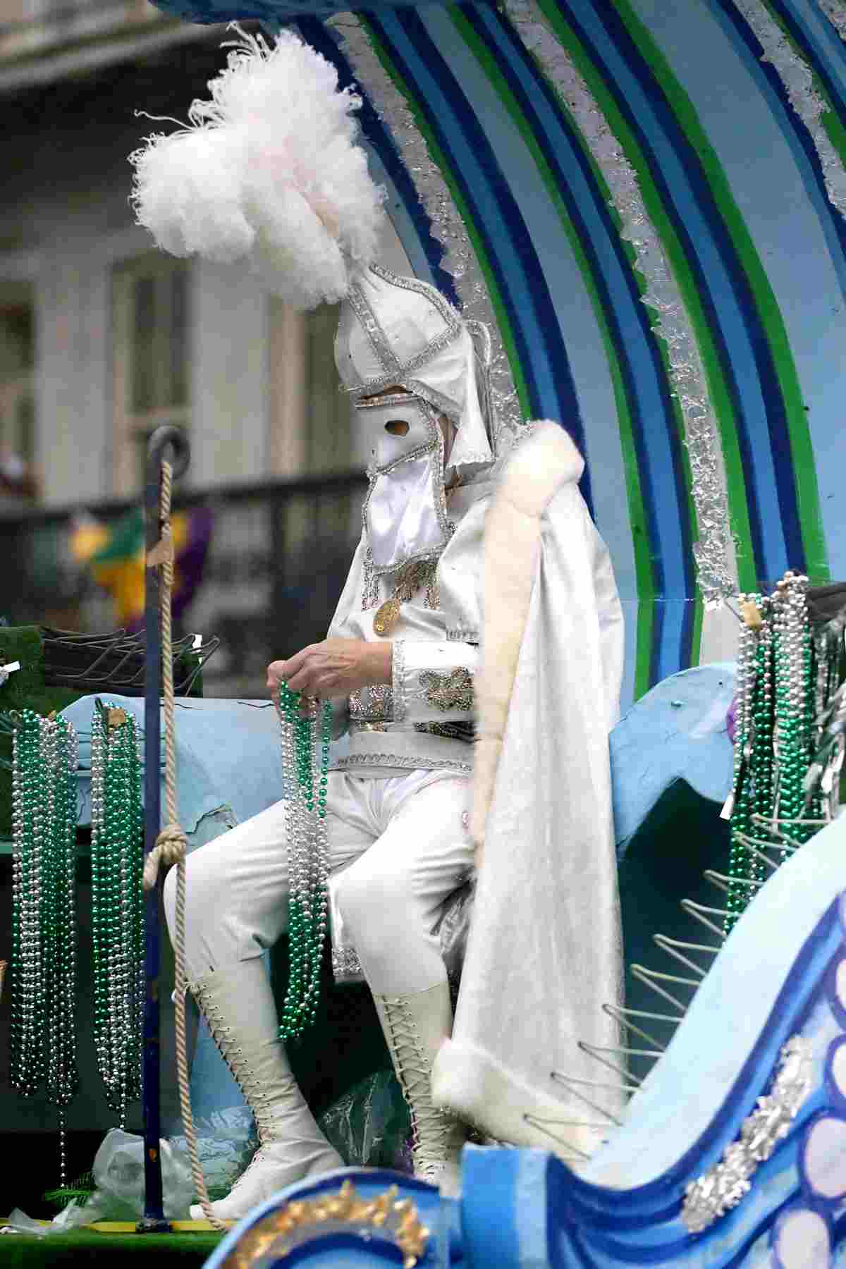 A Mardi Gras krewe member dressed in costume with a mask, sitting on a Mardi Gras parade float, holding beads that he is getting ready to throw.