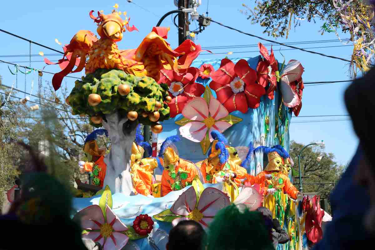 A Mardi Gras parade float with decorated with flowers, a tree, and a bird, with several people wearing Mardi Gras masks riding the float.