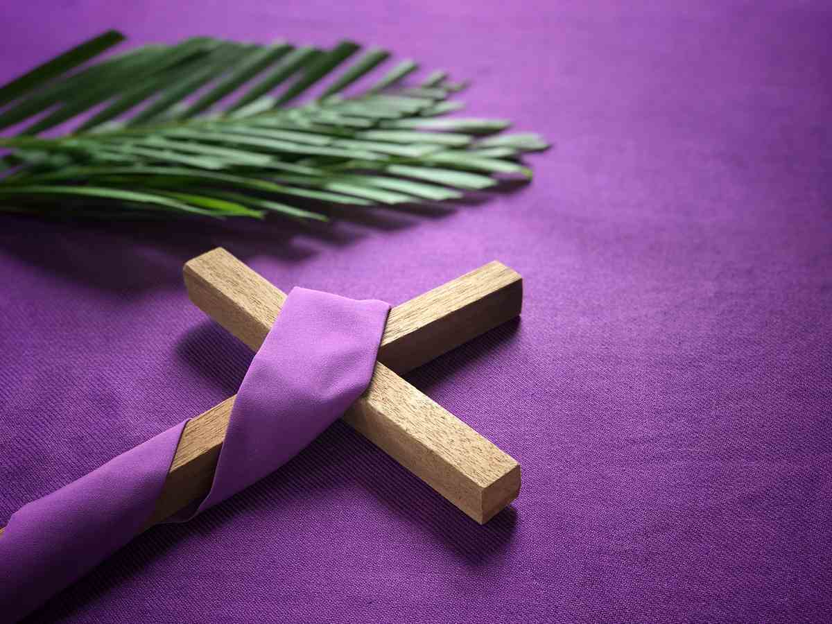 A purple background with a wooden cross wrapped in purple cloth next to palm leaves.