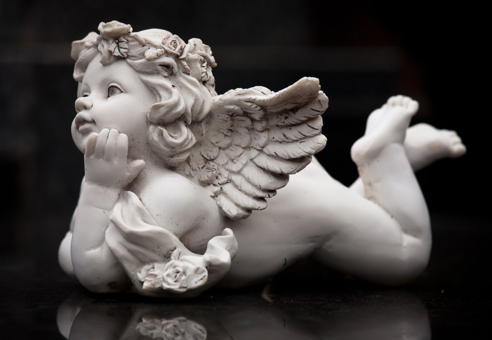 Baby Cupid statue laying down A carved stone statue of Cupid as a baby with wings, laying on his belly.