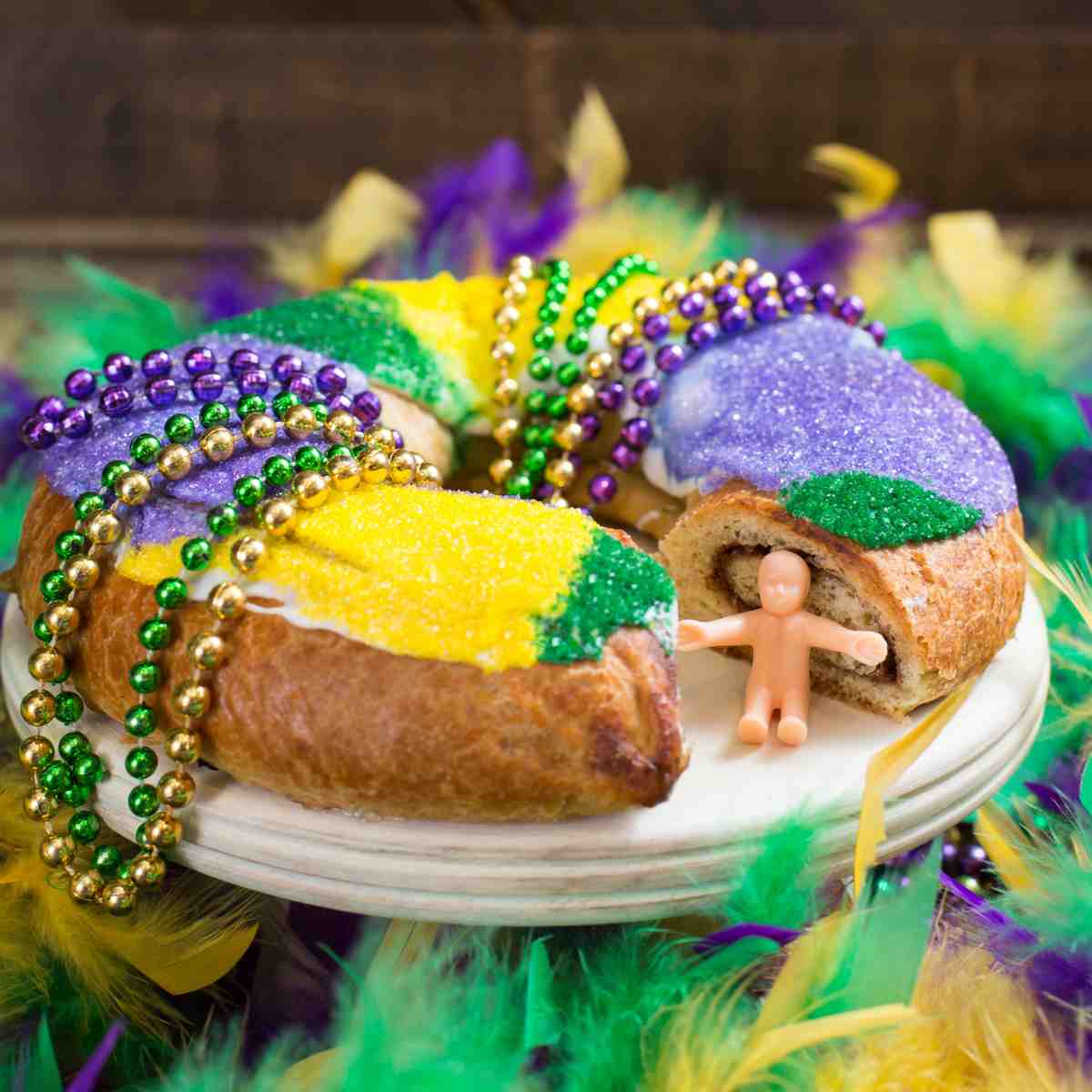 A king cake with one piece cut out on a cake stand draped with Mardi Gras beads, and with the plastic baby outside of the king cake.