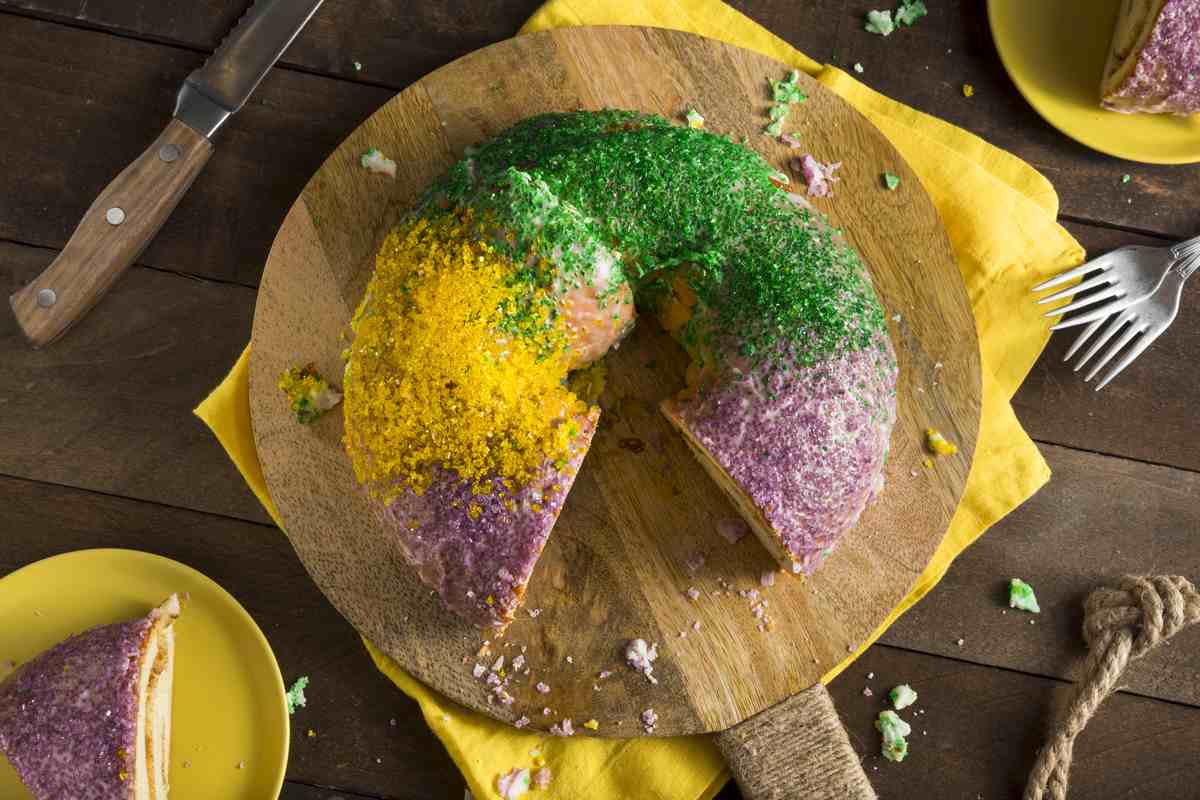 King cake A king cake with purple, green and gold sprinkles laying on a circular cutting board with a slice missing and a yellow towel in the frame.