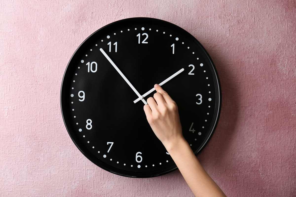 Clock at 2am A black clock on a lavender colored wall, with a hand reaching up to change to time for daylight saving time.