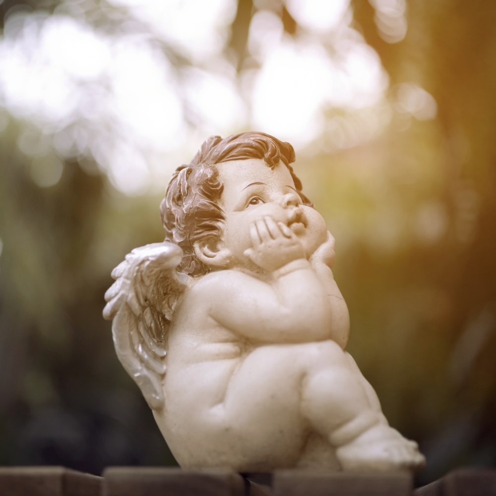 Baby Cupid statue A statue of a baby Cupid with his head in his hands, looking sad outside with blurred trees in the background.
