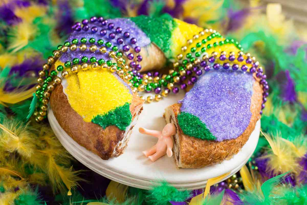 King cake, Mardi Gras beads and plastic Jesus A king cake with purple, gold and green sprinkles, draped with Mardi Gras beads and a baby from the king cake sitting next to the cake where a slice is missing.