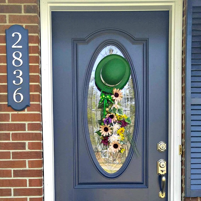 Leprechaun crafts A photo of a leprechaun craft which is a door swag made from a green leprechaun hat and seasonal flowers on a blue door of a home with a brick wall.