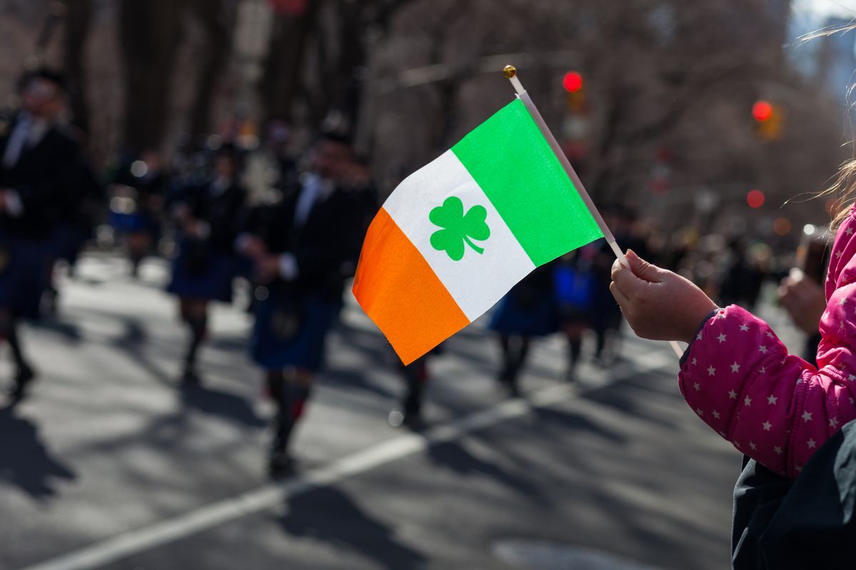 Parade and St. Patrick's Day flag A little girl's hand holding a small Irish flag with a shamrock on it during a St. Patrick's Day parade with people marching in the background.