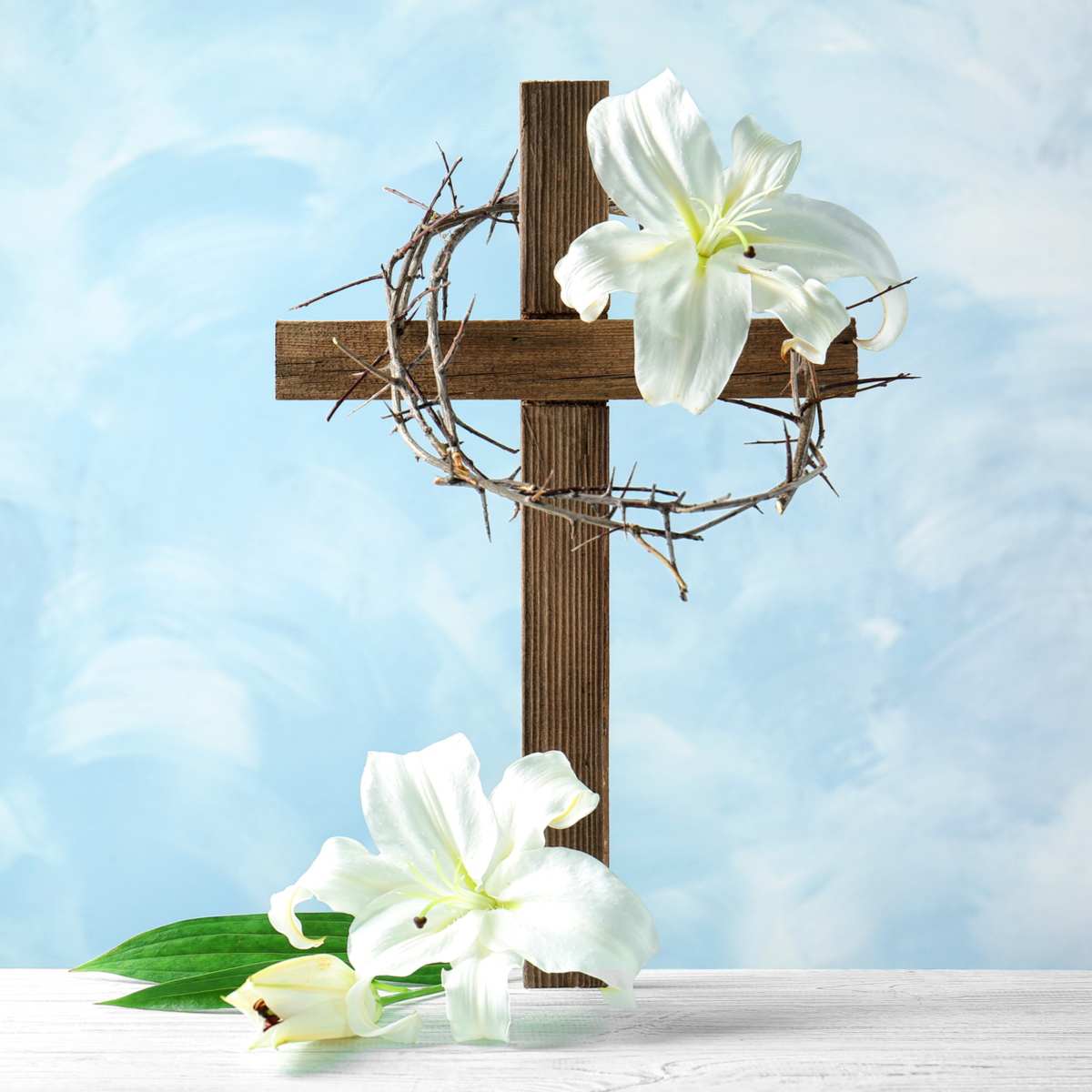 A cross with white Easter lilies at the bottom of it, and at the top right of it, with a crown of thorns draped around the top of the cross, all against a blue background.