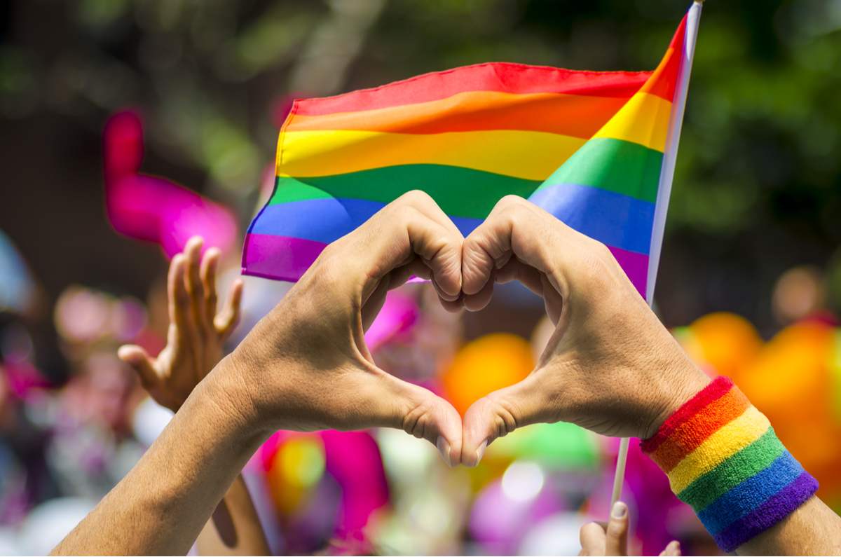 Pride flag and hands in a heart A pride flag waving during a parade with a person wearing a rainbow wrist band holding up their hand in the shape of a heart in the foreground. The perfect image for National Find a Rainbow Day.