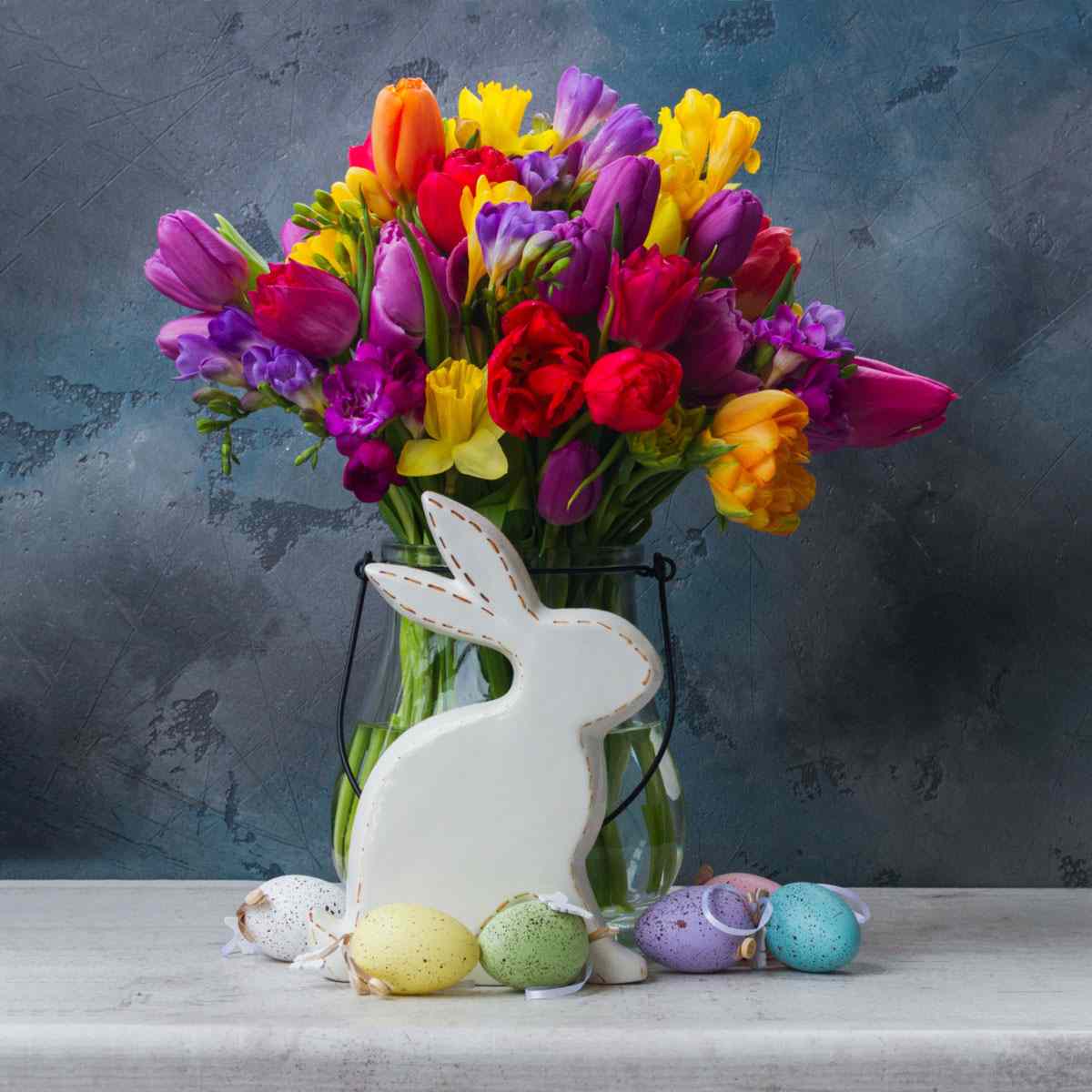 A bouquet of traditional Easter flowers in a clear vase with a white wooden Easter bunny in front of it, and pastel Easter eggs scattered around it.