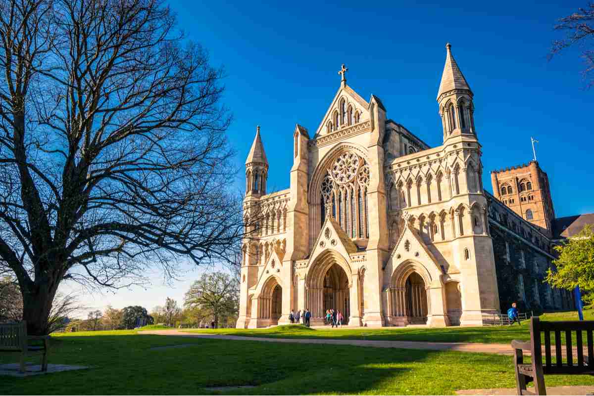 St Albans cathedral The origin location of the Alban bun, called St Albans Cathedral, against a bright blue sky, with green grass, the edge of a bench and tree without branches in the foreground of the photo.