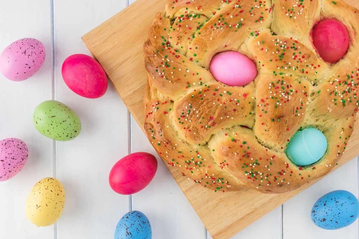 Pane di pasqua (Italian Easter bread) A pane di pasqua (Italian Easter bread) with dyed Eggs in it on a wooden cutting board, on top of a white wooden background with other pastel Easter eggs arranged around it.