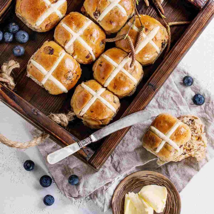 A wooden tray of hot cross buns, a ramekin of butter, a butter knife, and blueberries surrounding the tray.