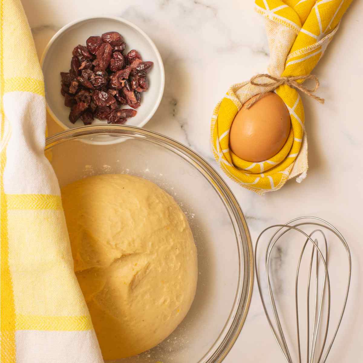 Hot cross bun ingredients The ingredients for hot cross buns on a beige table, including a bowl of raisins in the top left corner, an egg wrapped in a yellow dishcloth in the top right corner, a whisk in the bottom right corner, and a bowl of dough which is proofing in the bottom left corner.