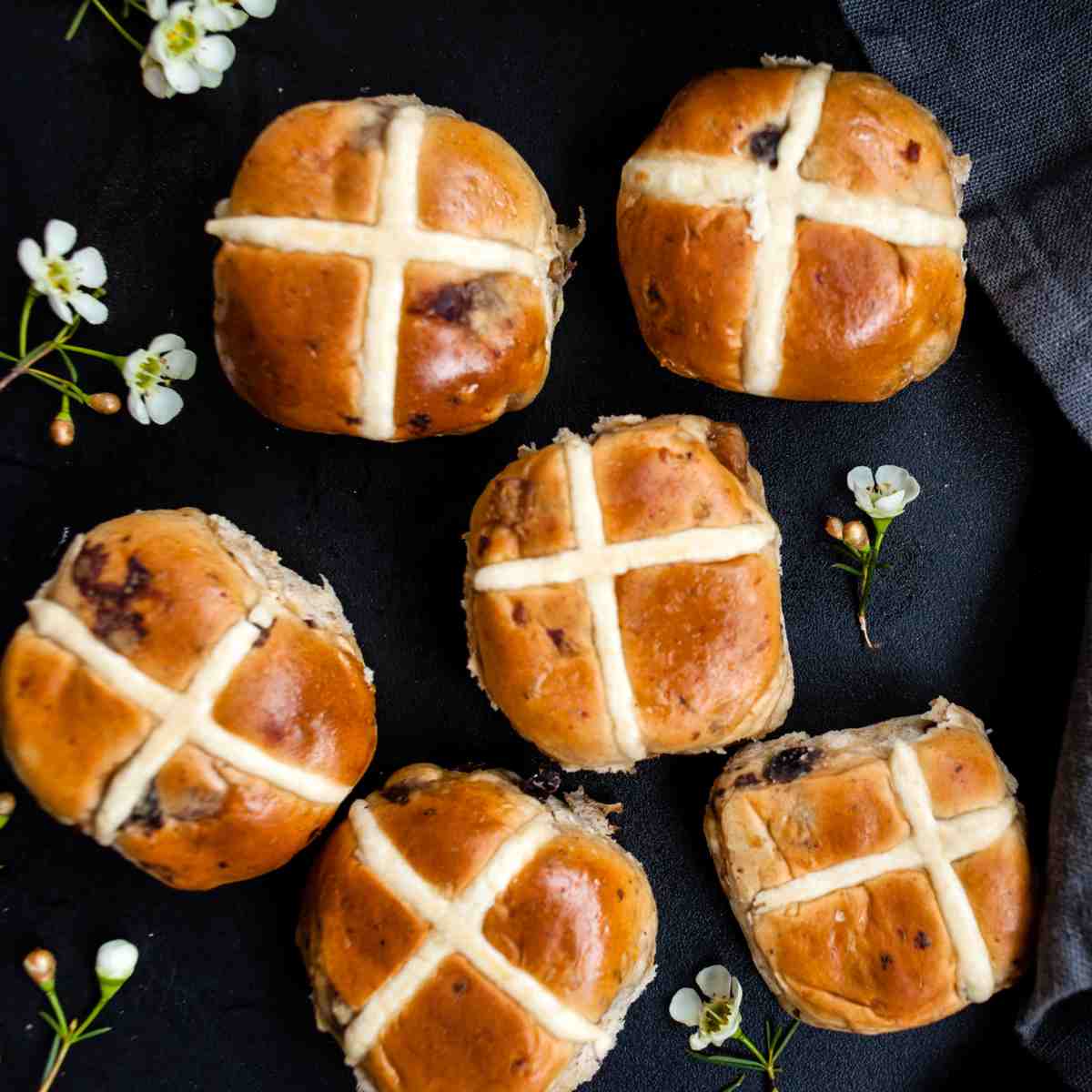 Hot cross buns and tiny flowers Six hot cross buns on a black napkin with tiny white flowers surrounding them, the cross prominently displayed on each of the Easter pastries symbolizing the religious hot cross buns history.
