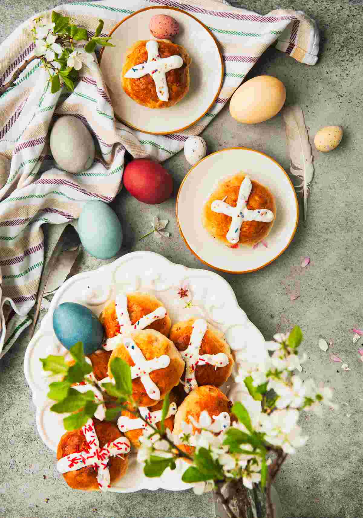 Easter themed hot cross buns Traditional hot cross buns at Easter on three separate plates, one plate containing multiple hot crossed buns, and the other two plates containing one of the Easter breads each, with Easter eggs, kitchen towels, and Easter flowers surrounding the Easter pastries.