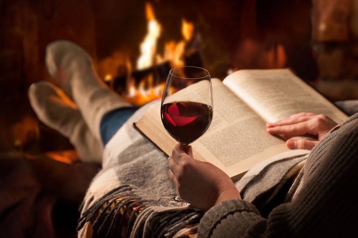 Wine and book near fire place A person wearing a sweater, jeans and cozy socks, holding an open book and a half full glass of red wine at the ideal red wine temperature in front of a fire.