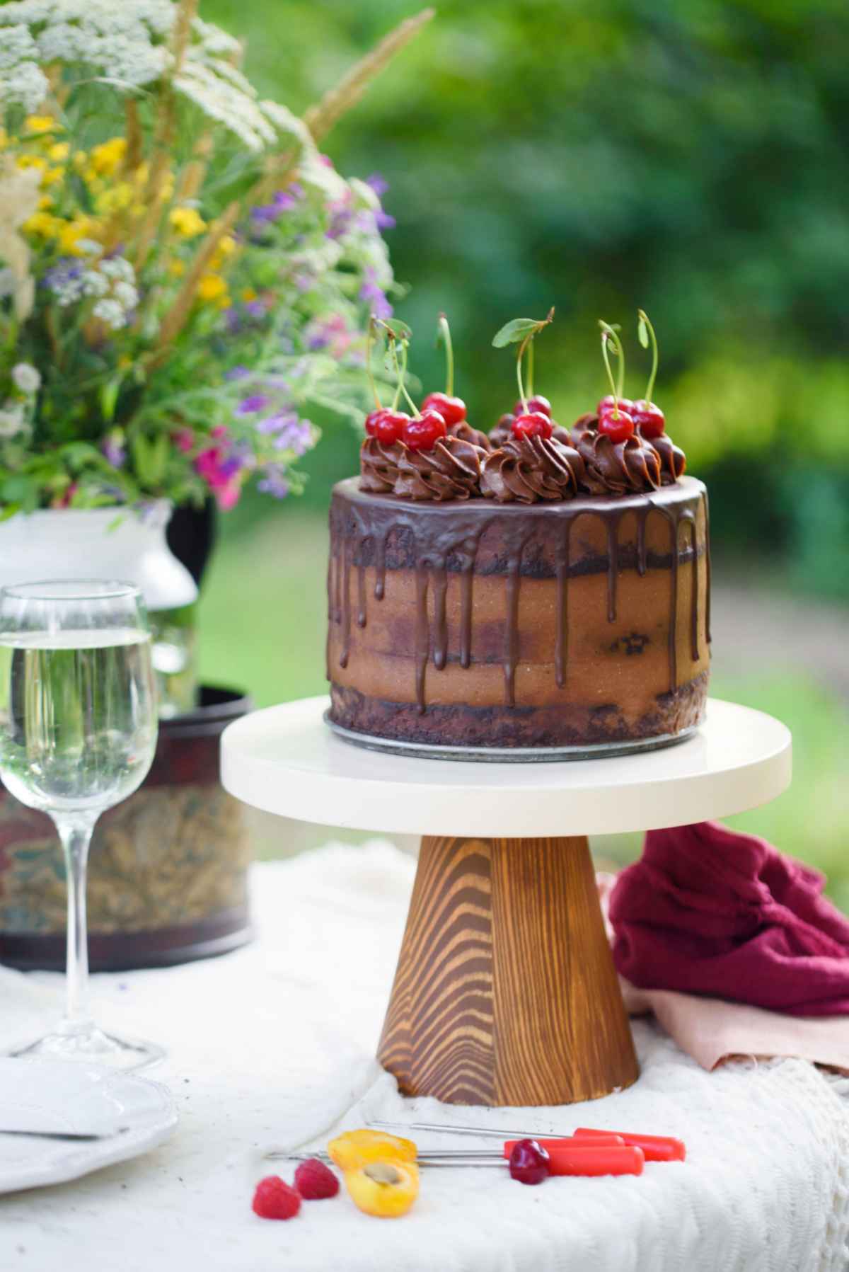 Chocolate cake on cake stand A german chocolate cake with cherries on top, sitting on a cake stand on a table outside in the summer next to a white wine glass.