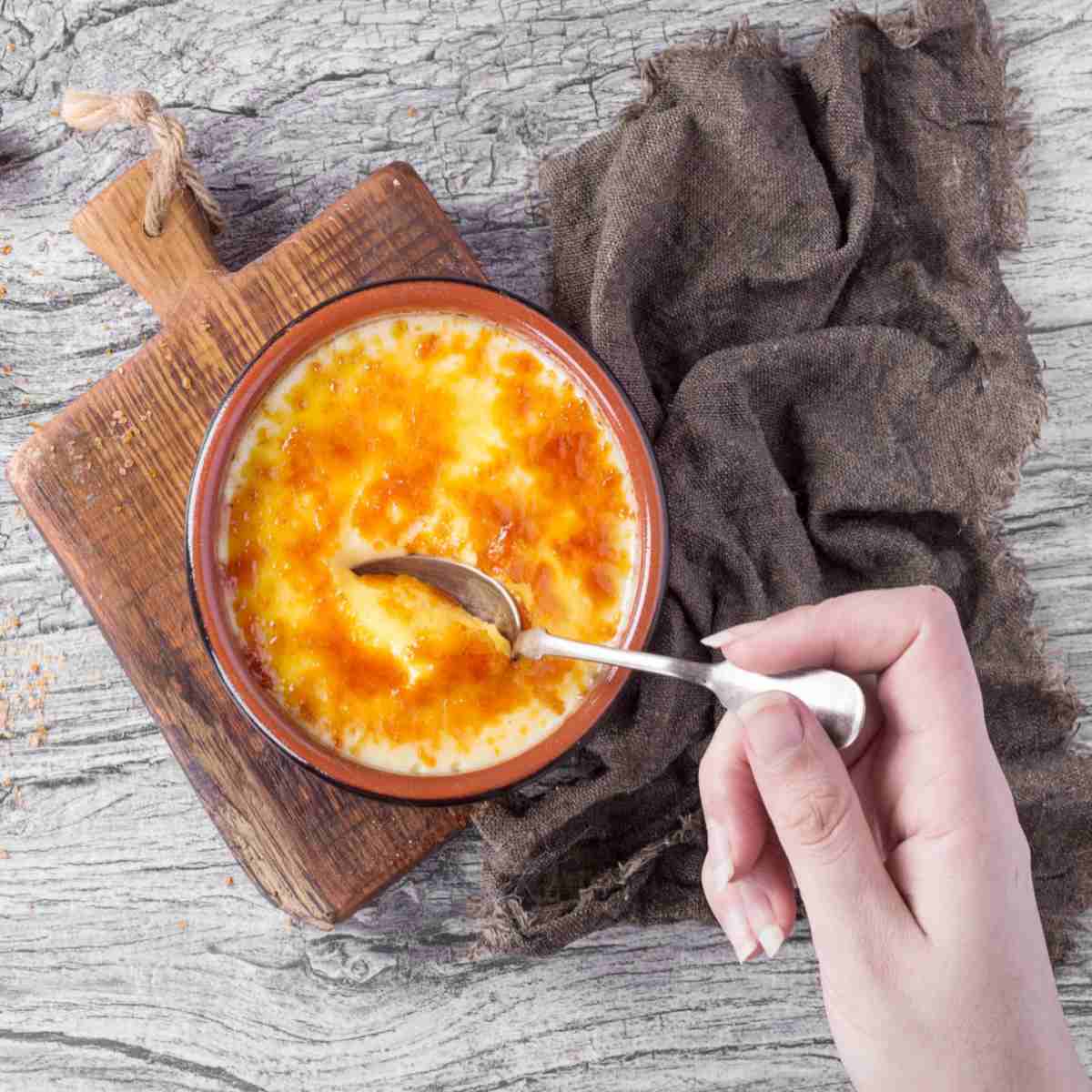 Creme brulee with spoon A woman's hand holding a spoon breaking the crust of a creme brulee on a cutting board with a brown towel beside it.