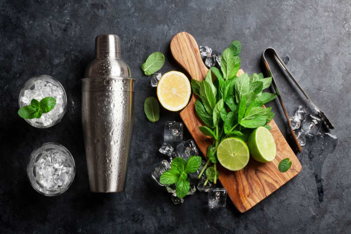 Mojito ingredients Mojito ingredients on a slate grey background, from left to right: ice in collins glasses, a cocktail shaker, a cutting board with limes and mint, and ice tongs.
