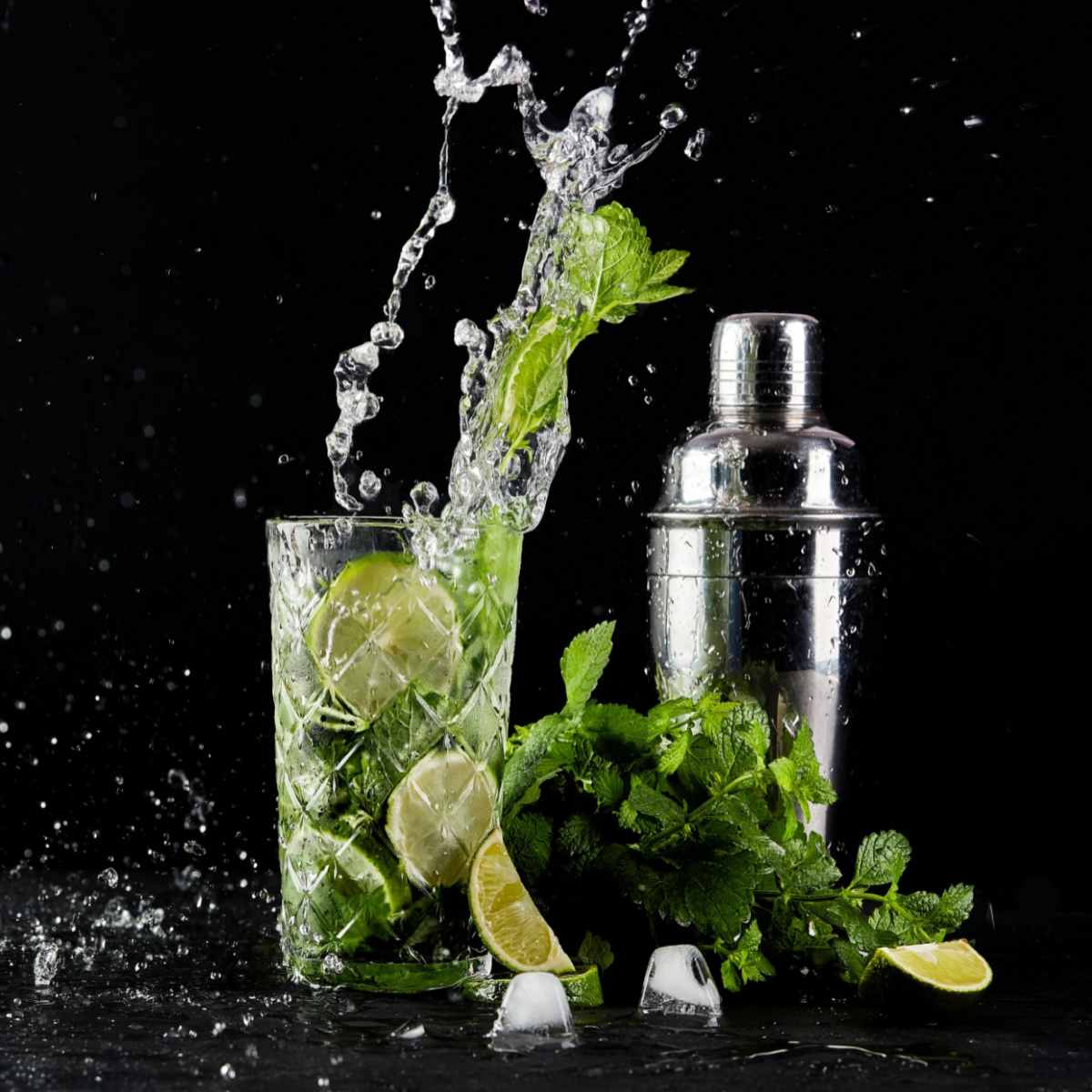 Mojito cocktail and shaker A mojito drink with ice splashing in it, next to a cocktail shaker, against a black backround.