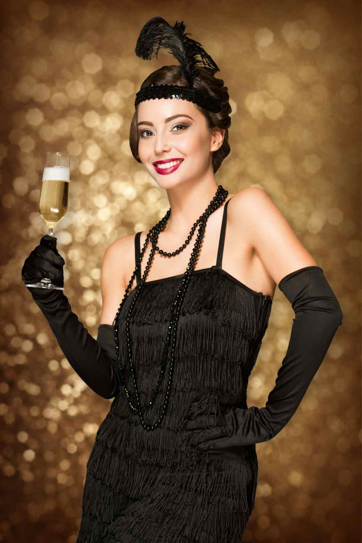 DIY flapper costume A woman wearing a DIY flapper costume, consisting of a black dress, black elbow length gloves, a black necklace and feathered headband, holding a glass of champagne against a background with golden bokeh.