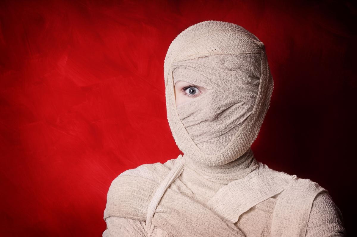 Mummy costume A diy mummy costume made of bandages, wrapped entirely around the person, with only her left eye visible, and she is standing in front of a red background.